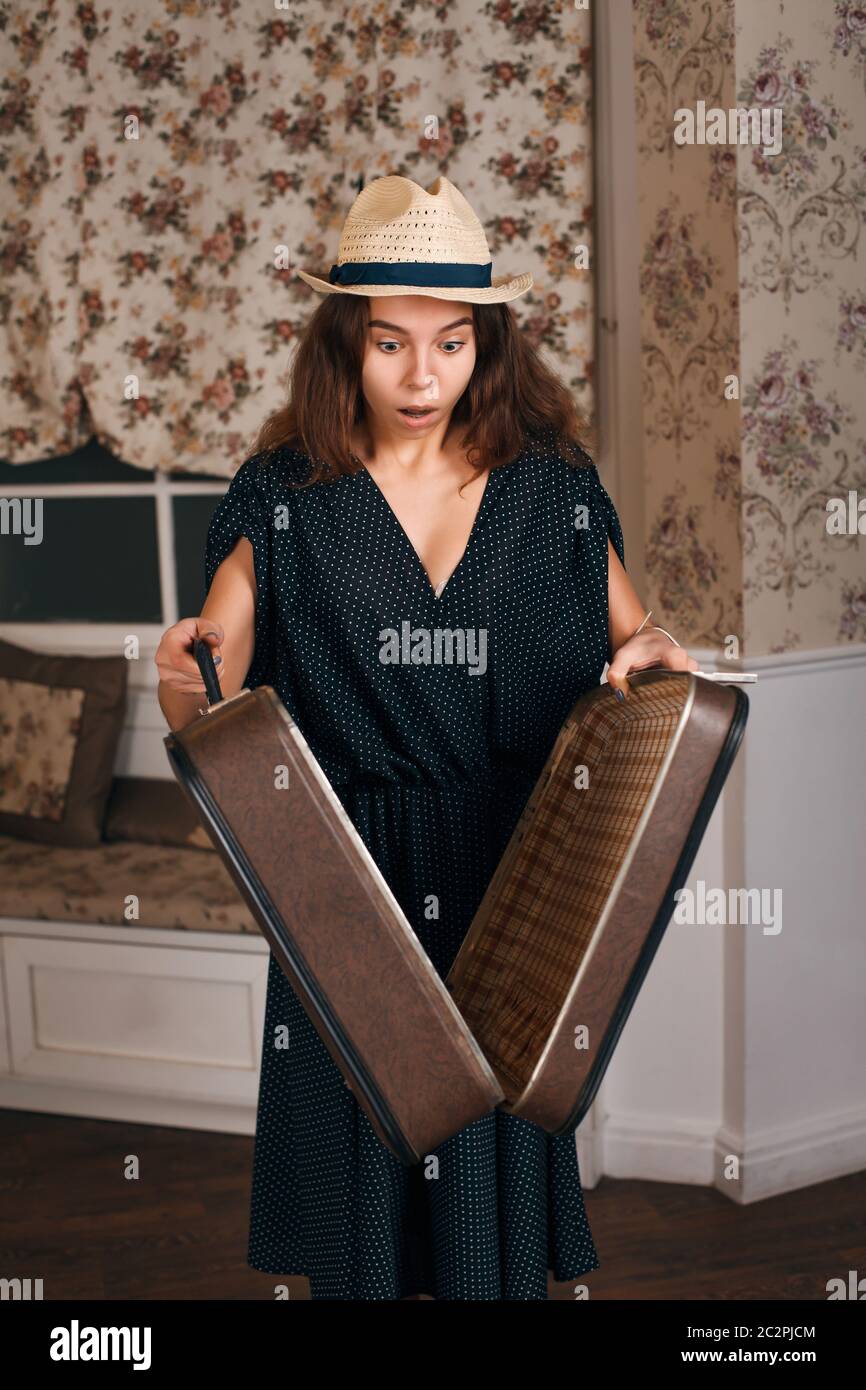Jeune femme en robe noire tenant une valise à moitié ouverte dans ses mains. Style vintage. Canapé et fenêtre avec rideaux sur l'arrière-plan. Banque D'Images
