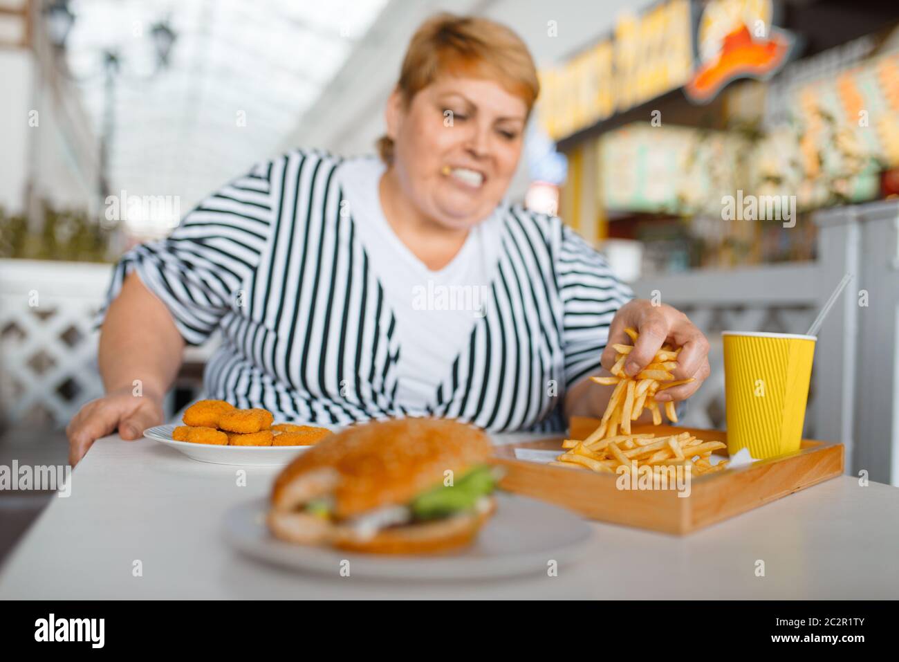 Grosse femme manger des calories des aliments dans le restaurant du centre commercial. L'excès de personne de sexe féminin à la table avec l'ordure dîner, problème d'obésité Banque D'Images