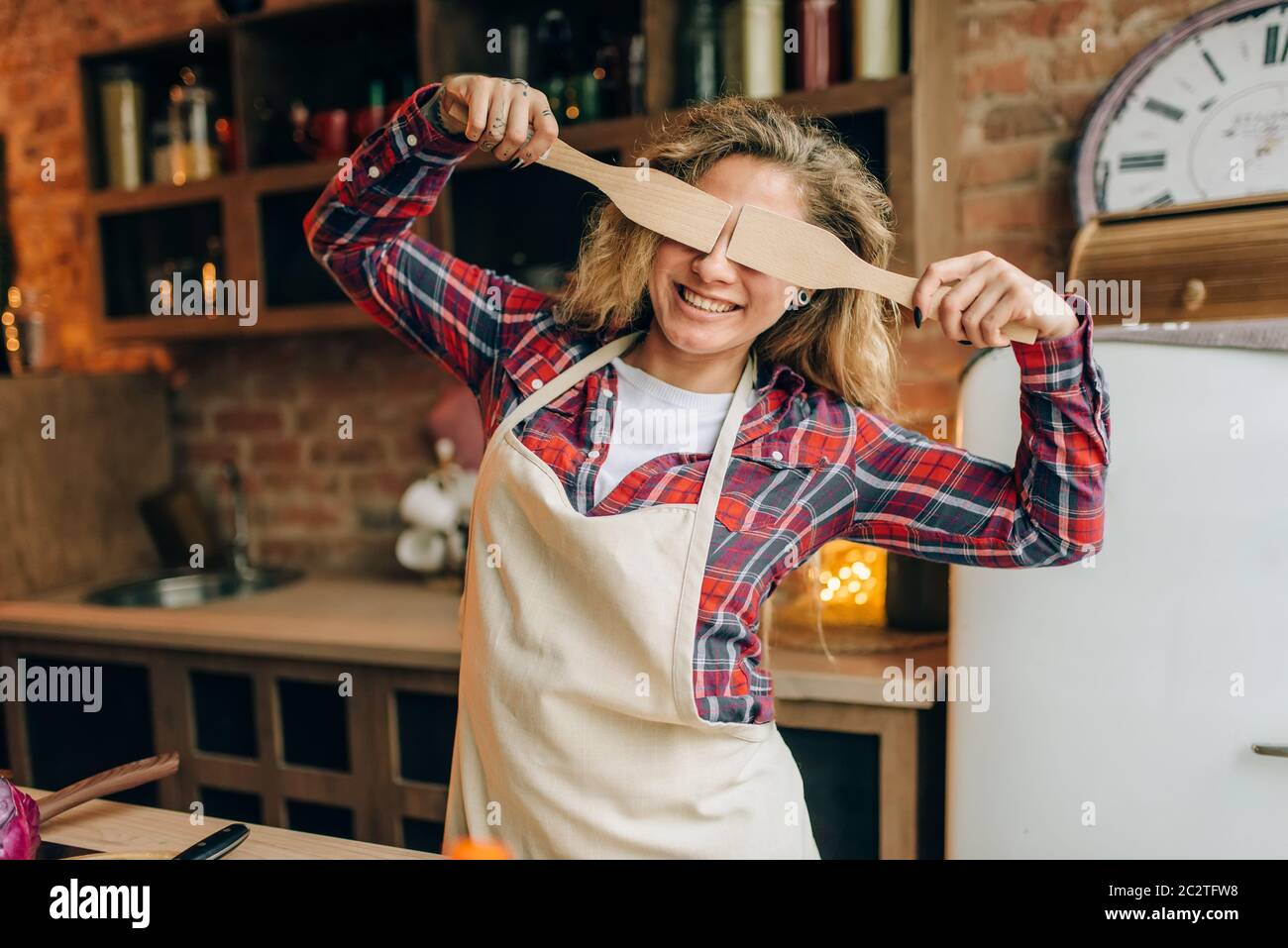 Femme au foyer ludique en tablier couvre ses yeux avec spatules en bois, cuisine intérieur sur arrière-plan. Heureux femme cuisinière faisant des choix de nourriture végétarienne, Banque D'Images