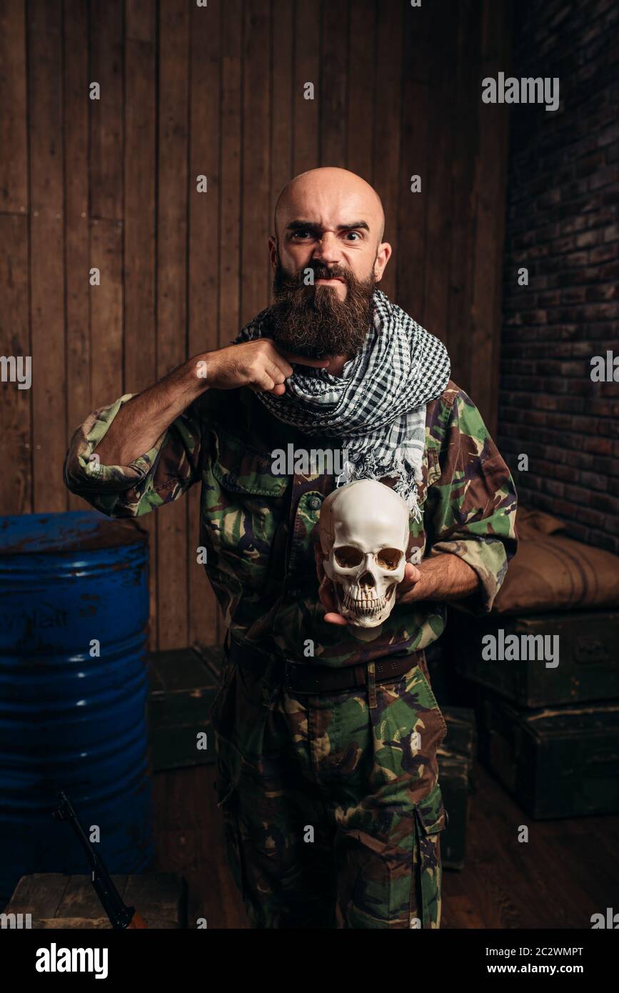 En uniforme terroriste détient crâne humain dans les mains, homme mojahed. Le terrorisme et la terreur horreur, soldat barbu en kaki de camouflage, de barils de carburant ou ch Banque D'Images