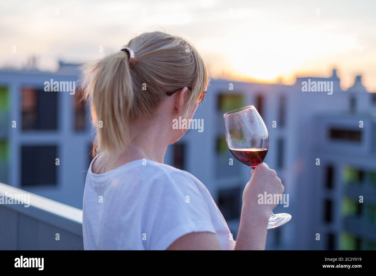 Blonde femme debout sur une terrasse et de boire un vin rouge au coucher du soleil Banque D'Images