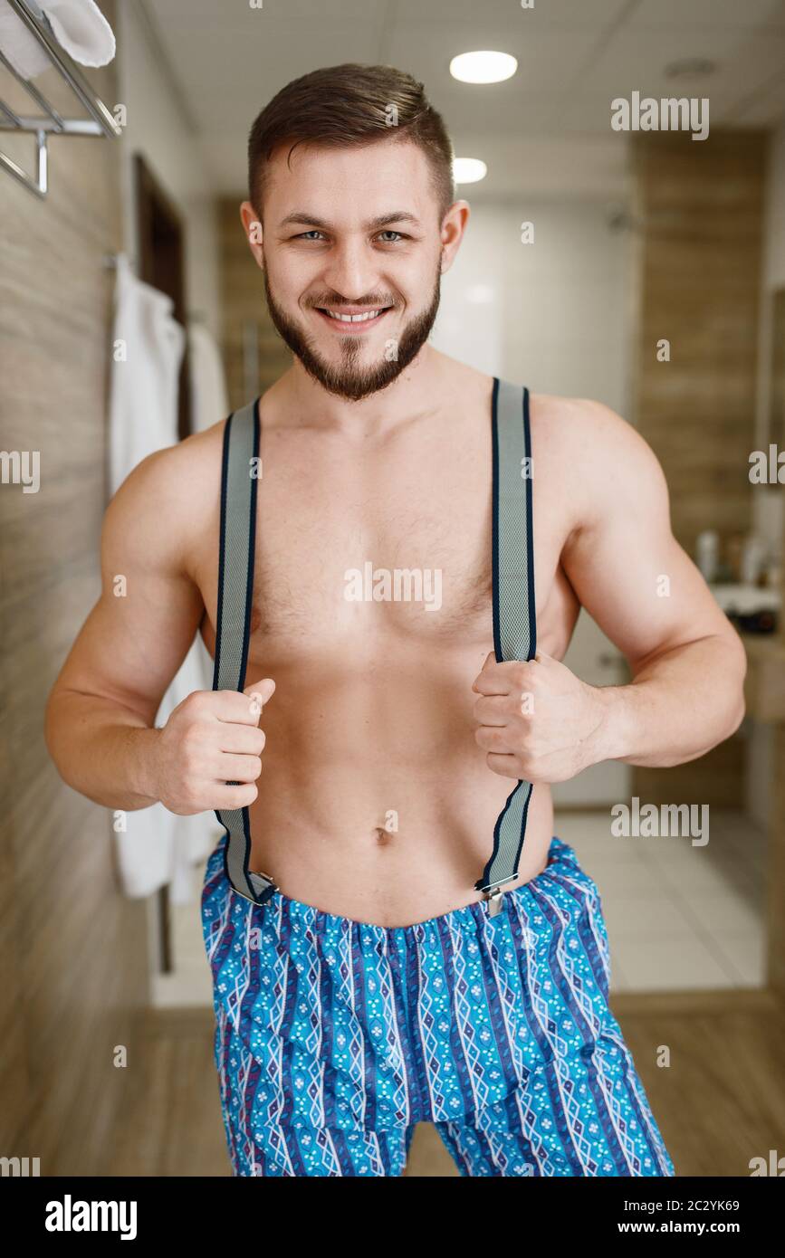 Homme barbu gai dans un pantalon avec bretelles pose dans la salle de  bains, préparation d'hygiène du matin. La personne masculine se prépare à  effectuer un traitement de la peau et du