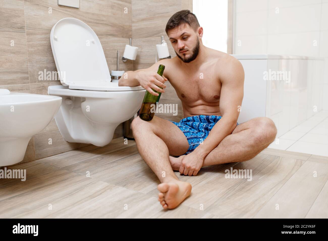 Homme de gueule de bois avec une bouteille de vin assis sur le sol près des  toilettes dans la salle de bains. Personne mâle hungover, mal de tête et  dépression, mauvais matin