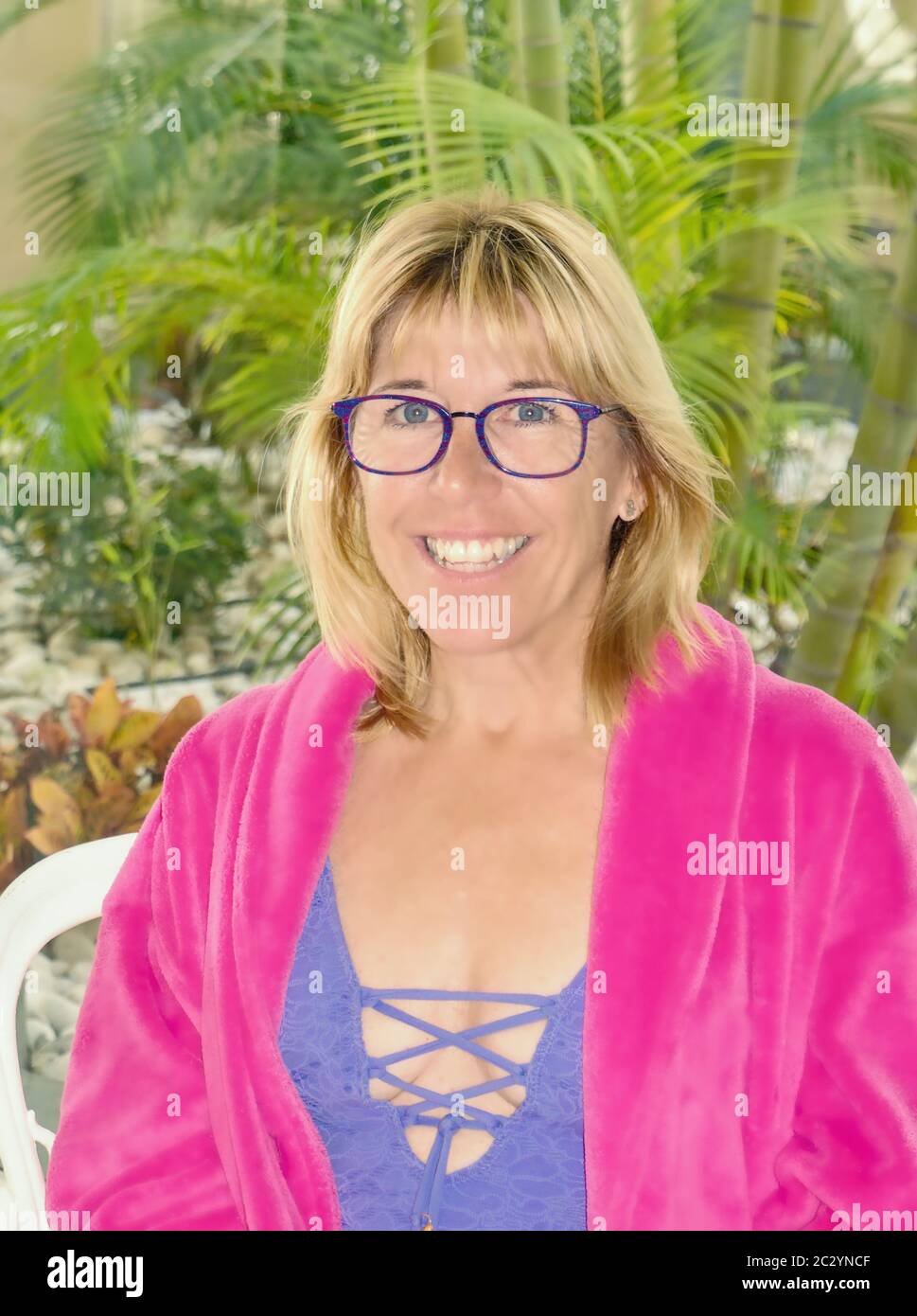 Une femme blonde aux yeux bleus et grand bleu lunettes dans la moitié portrait. Il est vêtu d'un peignoir moelleux ouvert rose, sous le maillot bleu. Pal vert Banque D'Images
