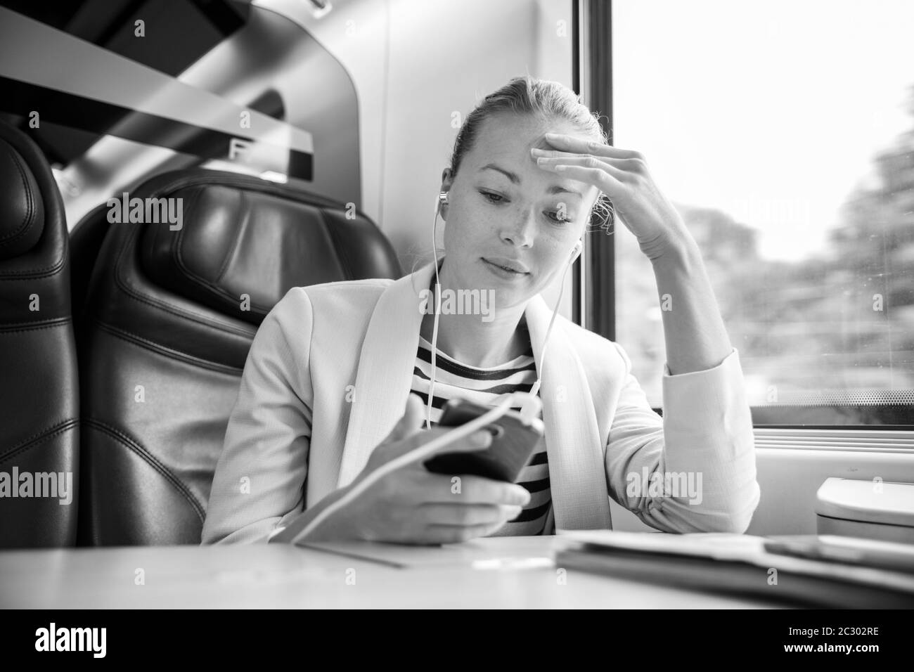 Femme d'affaires caucasienne communiquant sur un téléphone portable en utilisant un casque pendant son voyage en train dans un siège de classe affaires. Image en noir et blanc. Banque D'Images