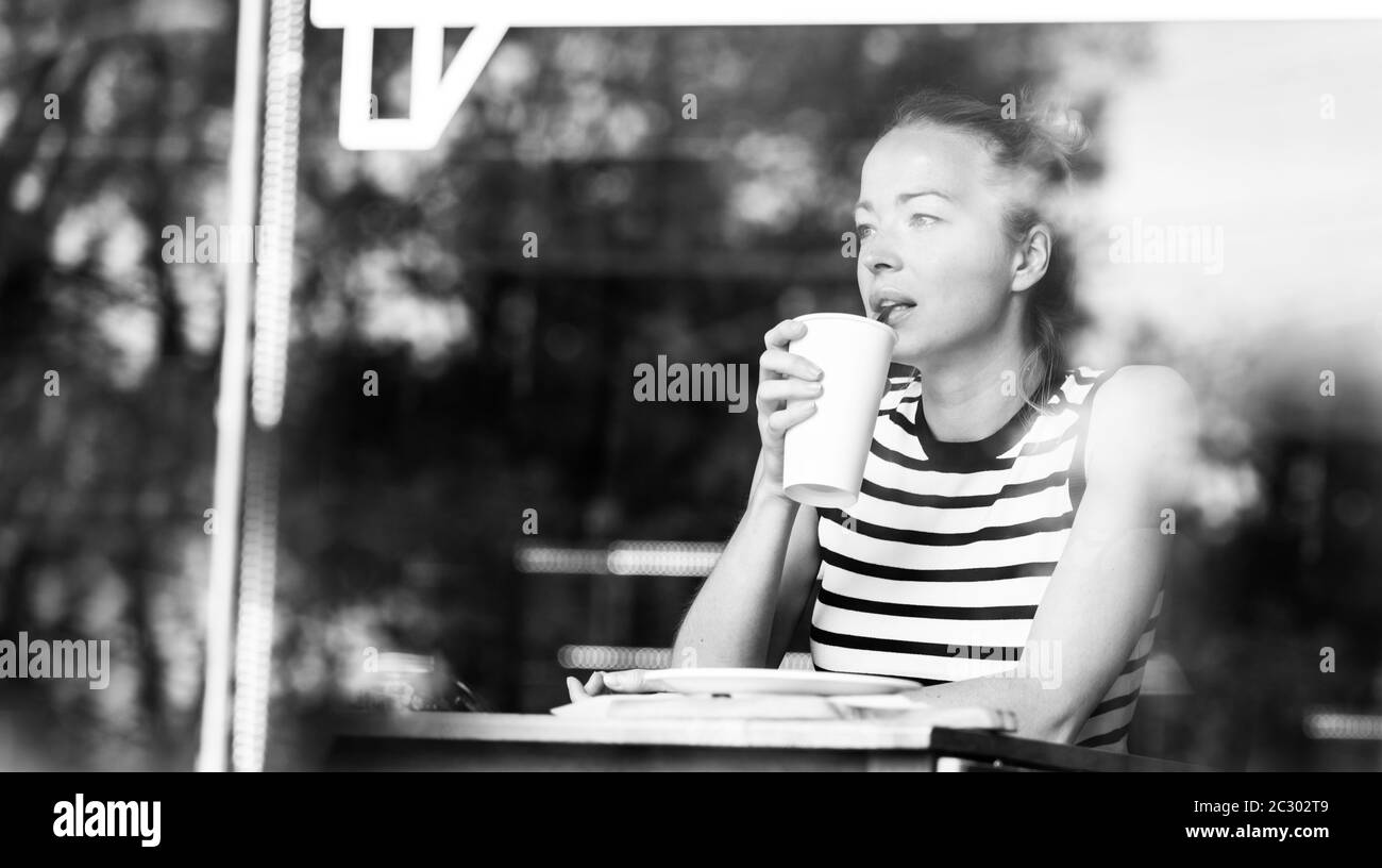 Jeune femme caucasienne assise seule dans un café, buvant du café américain, les gens regardant, regardant avec attention par la fenêtre du café. BLAC Banque D'Images