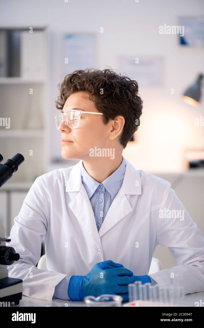 Virologue pensive et pratique aux cheveux bouclés en blouse de laboratoire et gants assis au bureau dans un laboratoire moderne et regardant loin Banque D'Images