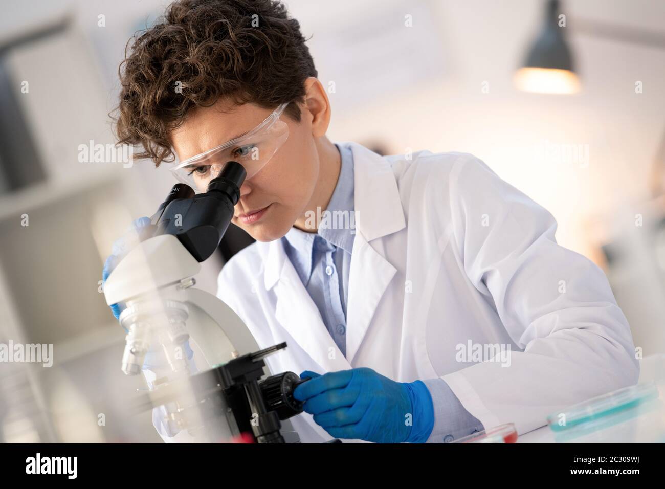 Chercheur concentré à poil dur de la société pharmaceutique tournant le bouton tout en ajustant le microscope en laboratoire Banque D'Images