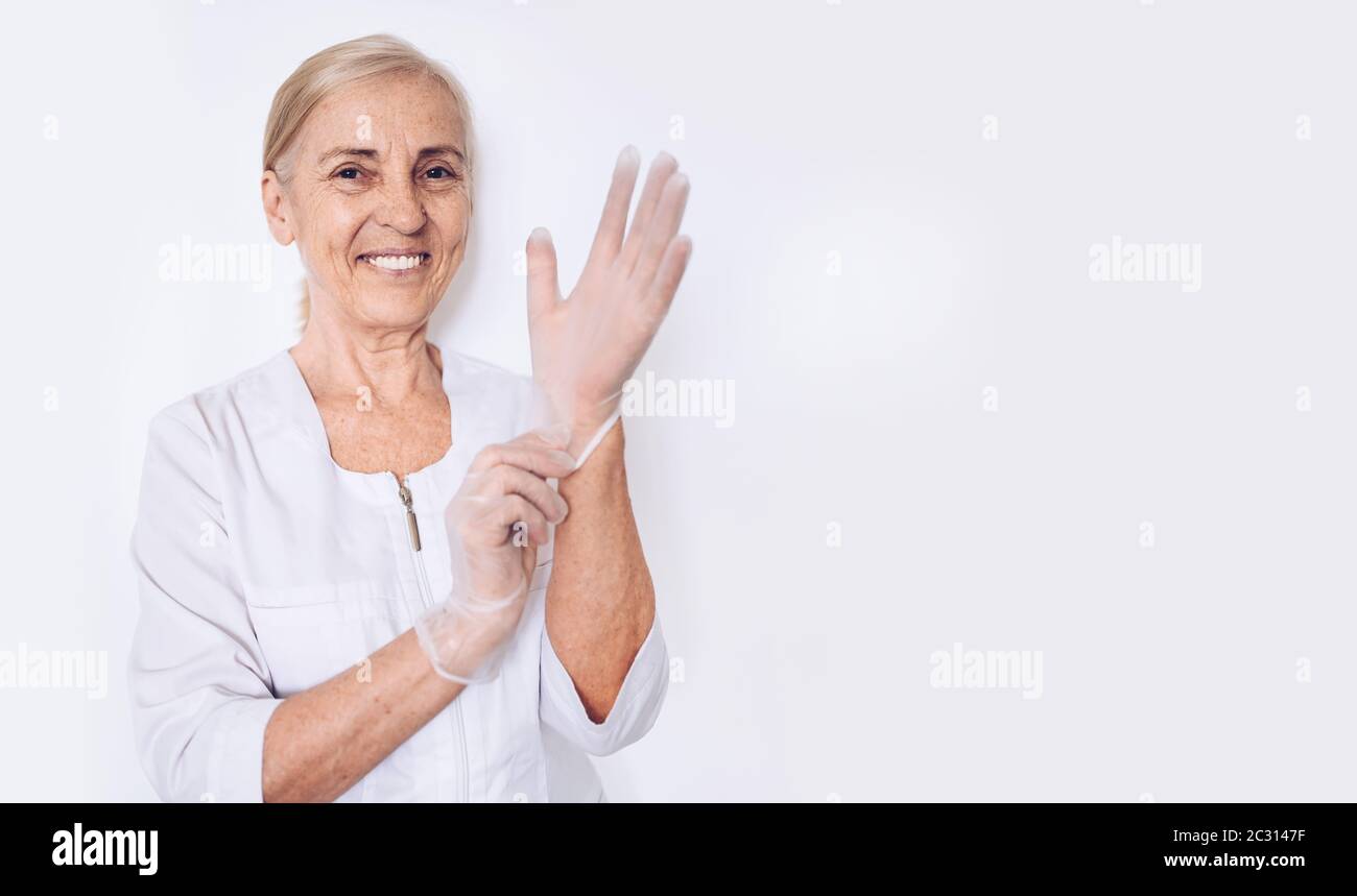 Femme âgée souriante femme mature médecin ou infirmière dans un manteau médical blanc met des gants portant un équipement de protection individuelle isolé. Santé et Banque D'Images