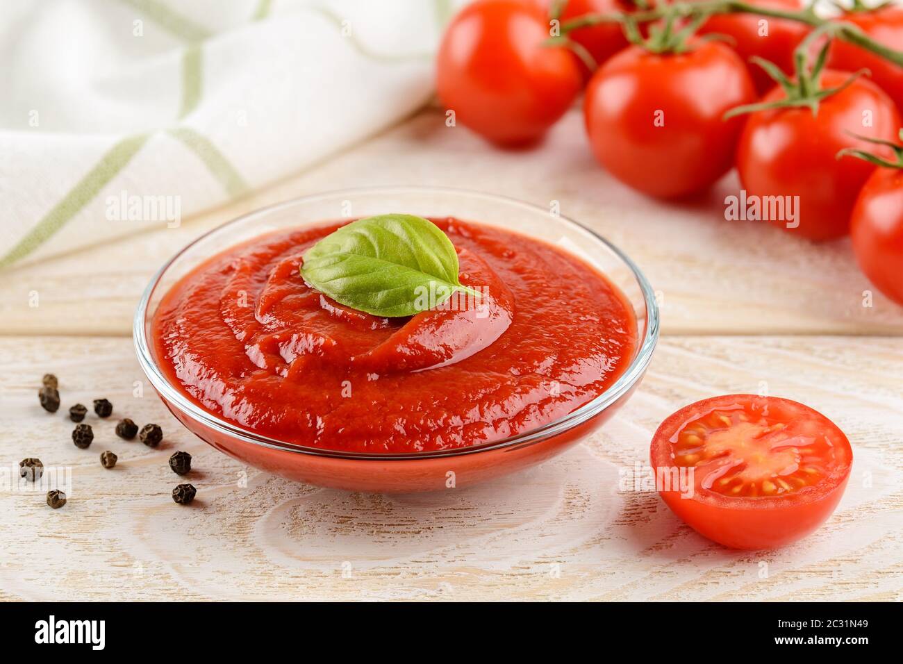 Ketchup de tomate épais et savoureux dans un bol en verre contre les tomates cerises rouges mûres sur une branche sur une table en bois blanc. Épices, assaisonnements et sauces. Gros plan. Banque D'Images