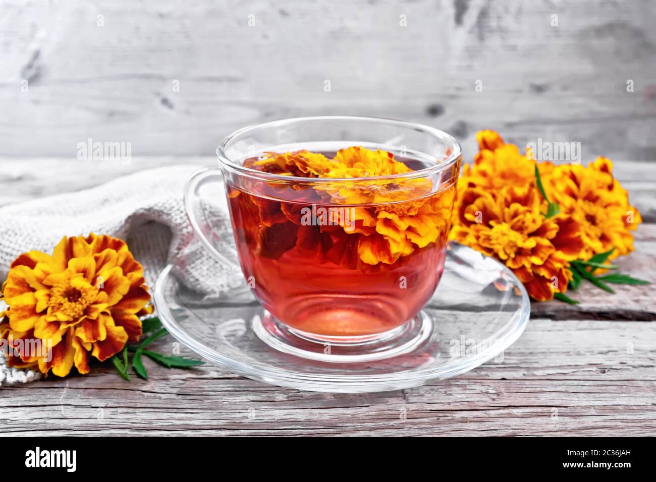 Thé aux herbes Marigold dans une tasse en verre et soucoupe, fleurs fraîches, serviette en toile de fond d'un vieux panneau en bois Banque D'Images