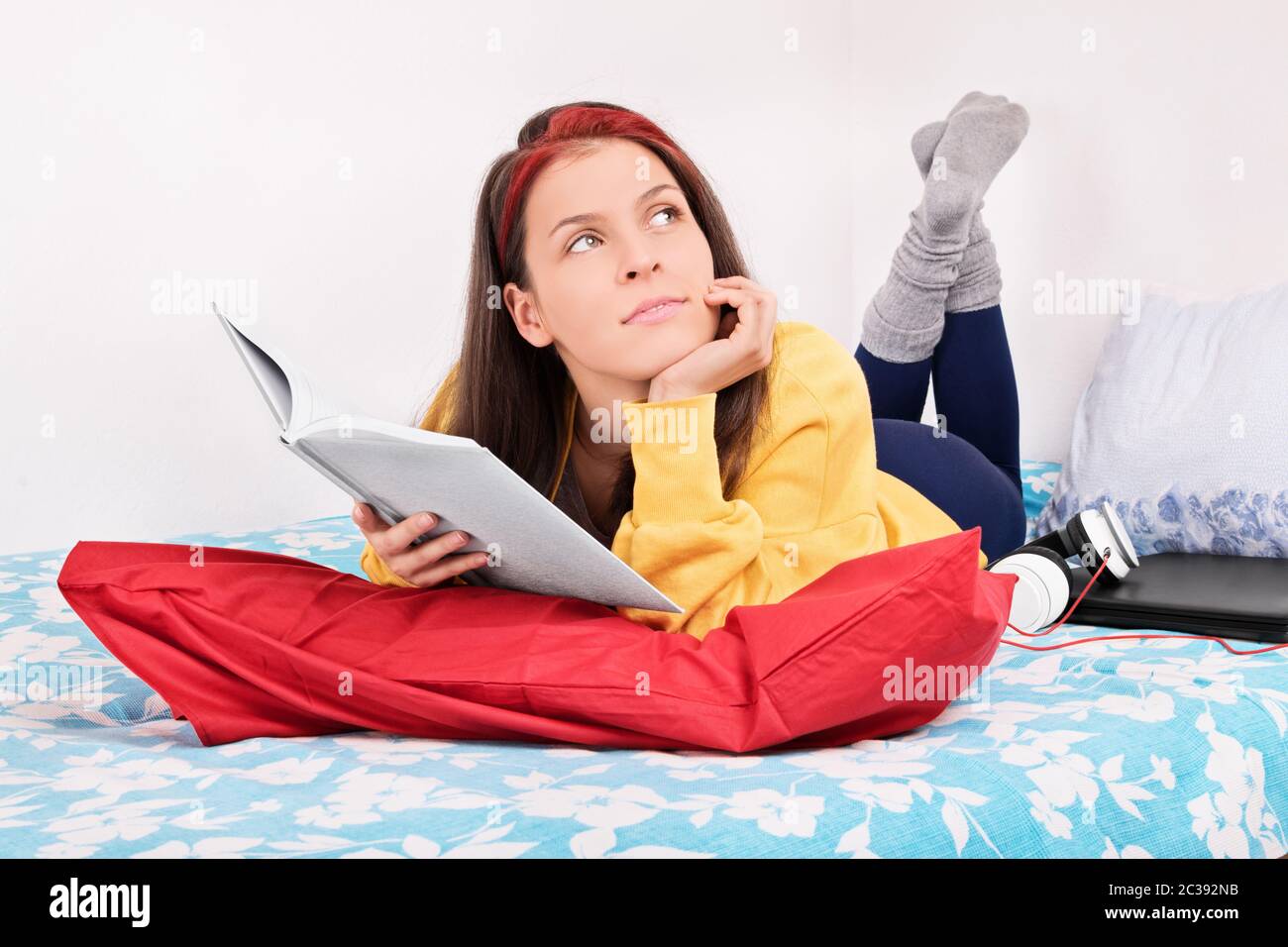 Belle jeune fille dans sa chambre tenant un livre, regardant à côté, pensant, se demandant, fantastizing tout en étant couché dans le lit à côté de l'ordinateur portable et du casque Banque D'Images