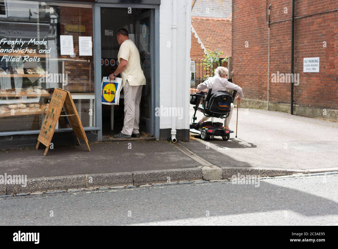 Homme achetant dans une boulangerie de rue haute par le biais d'un écran de protection en plastique à la porte du magasin tandis que la femme âgée dans un fauteuil roulant attend à proximité, Royaume-Uni Banque D'Images
