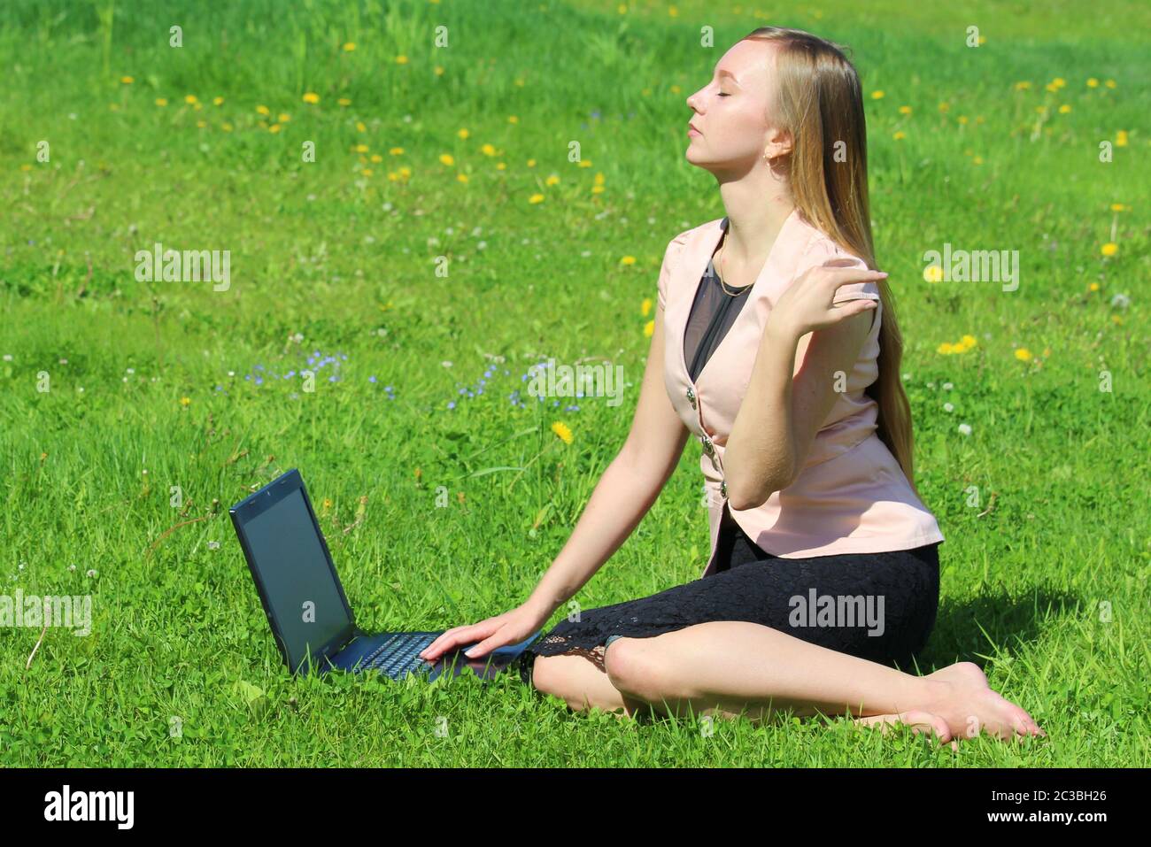 Une belle jeune fille blanche dans une veste rose et une jupe noire et avec de longs cheveux assis sur l'herbe verte, sur la pelouse et travaillant derrière un ordinateur portable noir. Banque D'Images