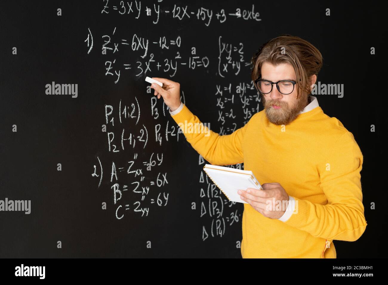 Professeur sérieux avec copybook pointant à l'équation sur le tableau noir avec morceau de craie tout en expliquant aux élèves en ligne comment la résoudre Banque D'Images