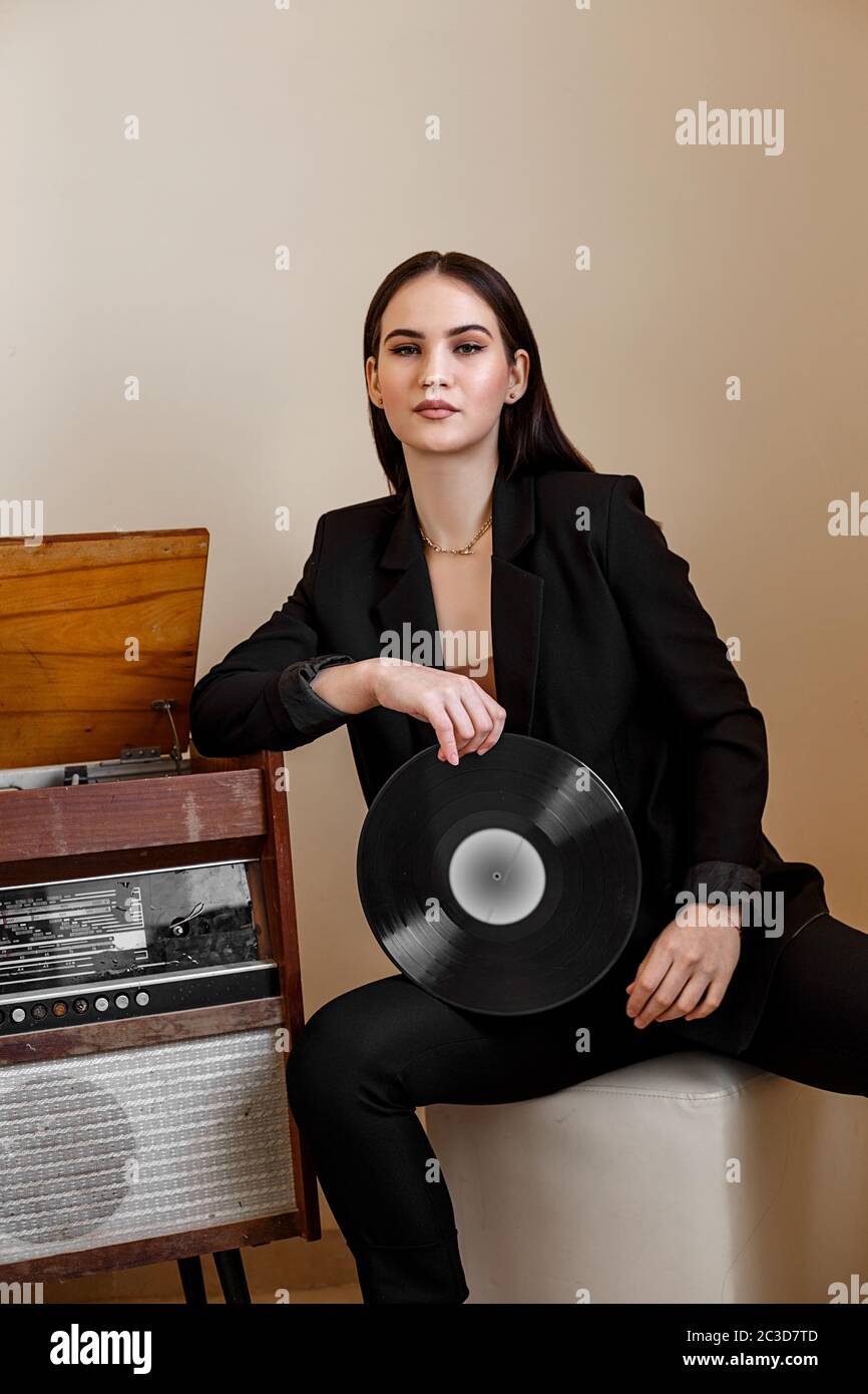 Une belle jeune femme caucasienne dans un pantailleur noir et des sandales noires pose à côté d'un ancien joueur de disque avec un gramophone Banque D'Images