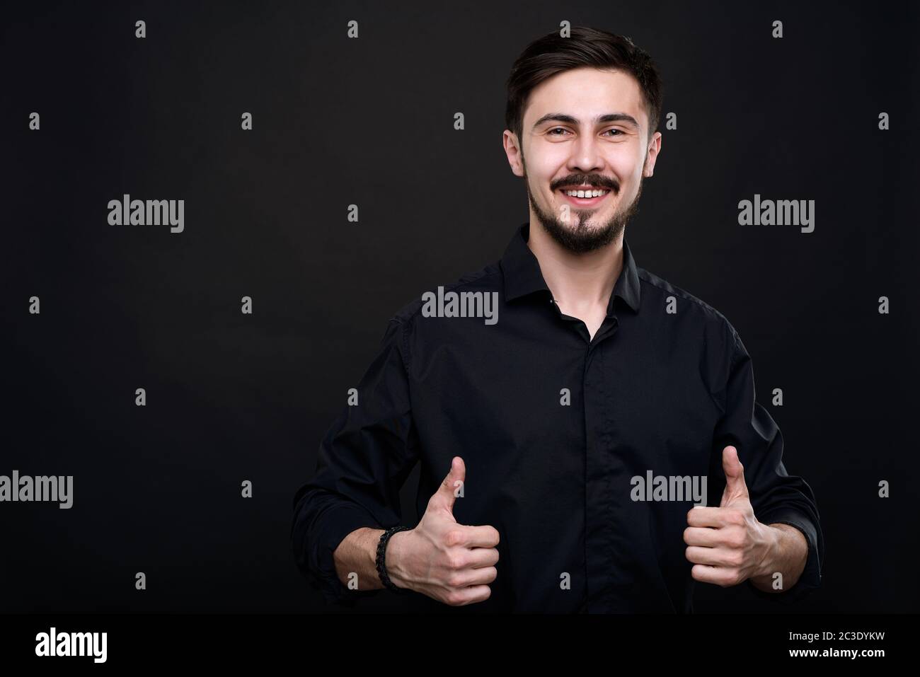 Portrait d'un jeune homme excité debout sur fond noir et montrant le pouce vers le haut comme symbole de qualité Banque D'Images