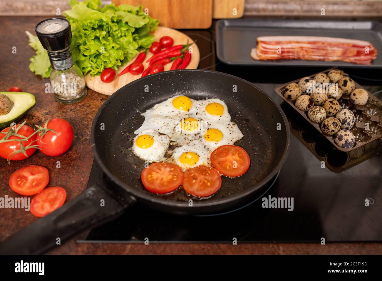 Plusieurs œufs de caille frits et tranches de tomates arrosées de poivre noir moulu sur une poêle entourée de légumes frais Banque D'Images