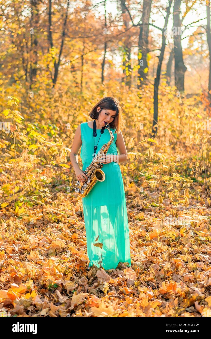 jolie fille avec cheveux noirs dans une robe bleue joue le saxophone alto au milieu du feuillage jaune dans un parc en automne Banque D'Images
