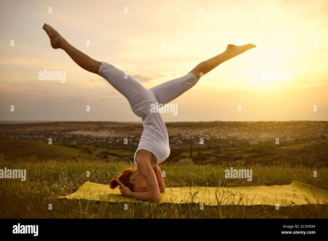 Une femme en vêtements de sport blancs pratique le balsa de yoga debout sur la tête sur l'herbe à l'extérieur au coucher du soleil. Banque D'Images