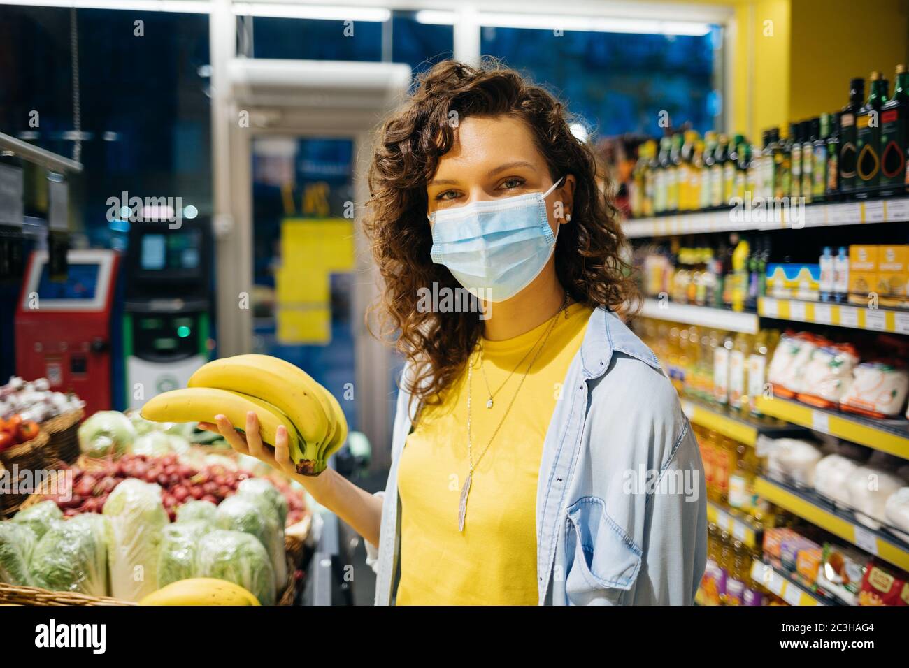 Jeune femme portant un masque de protection médical debout dans un magasin d'épicerie tenant un bouquet de bananes regardant l'appareil photo. Banque D'Images