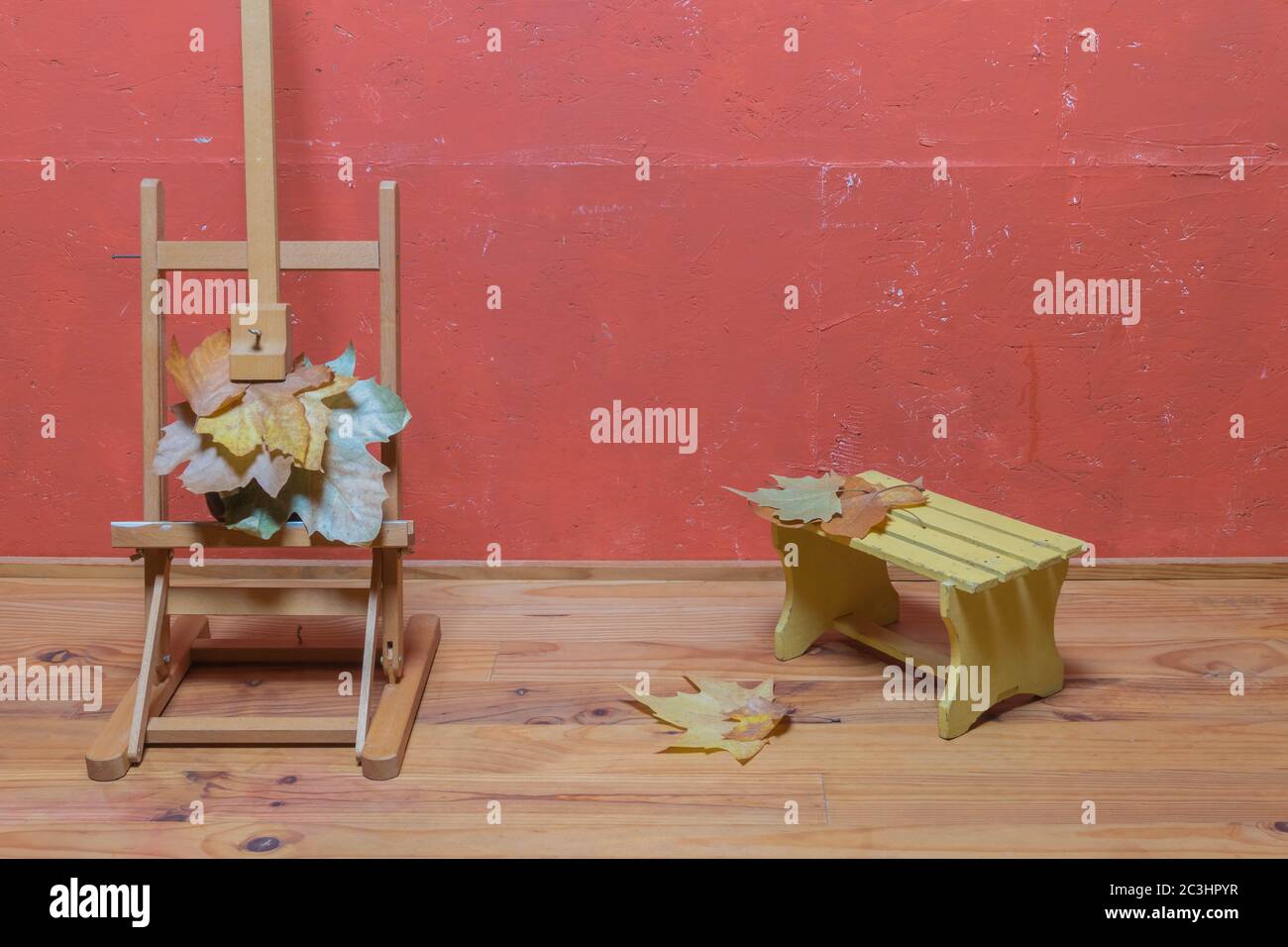 Support traditionnel en chevalet avec feuilles séchées, petit banc en bois, feuilles séchées sur parquet et mur peint en rouge en studio. Décoration d'automne. Banque D'Images