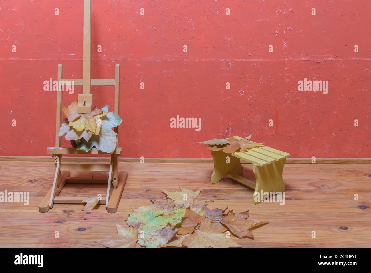 Support traditionnel en chevalet avec feuilles séchées, petit banc en bois, feuilles séchées sur parquet et mur peint en rouge en studio. Décoration d'automne. Banque D'Images