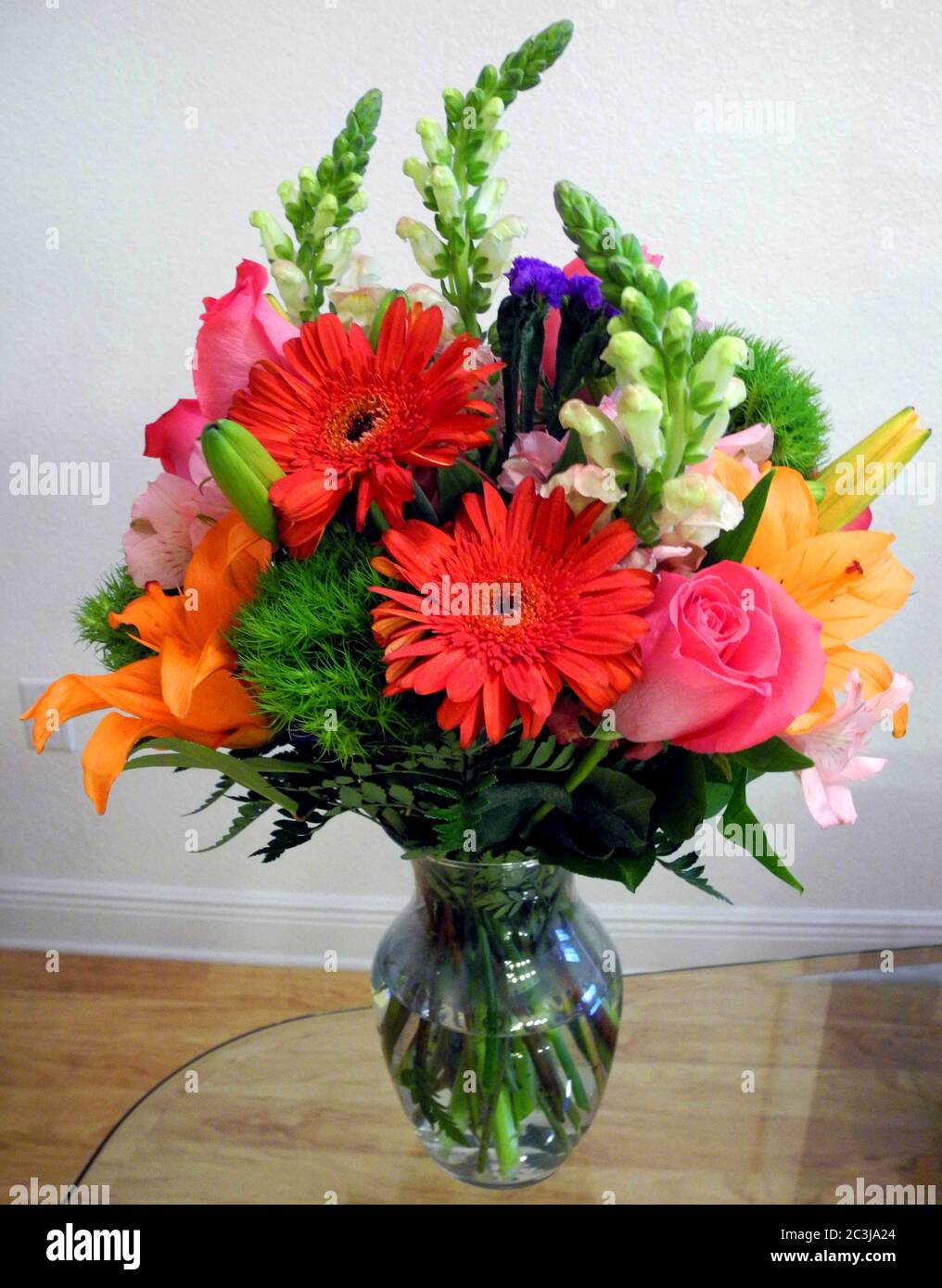 Un bouquet de fleurs coupées colorées de plusieurs variétés est présenté dans un vase en verre transparent sur une table recouverte de verre. Qu'ils soient cultivés dans un jardin à la maison ou achetés auprès d'un fleuriste, les jolies fleurs dureront plus longtemps si elles sont correctement préparées et soignées. Commencez par vous assurer que les tiges de fleurs sont vertes et lisses, pas minces. Avant de les mettre dans un vase, faites une coupe fraîche d'un pouce ou deux vers le haut de la tige et retirez toutes les feuilles qui seront sous la surface de l'eau. Placer les tiges dans de l'eau douce et ajouter un paquet de nourriture de fleur. Changez l'eau tous les deux jours, et faites une nouvelle coupe des tiges. Banque D'Images