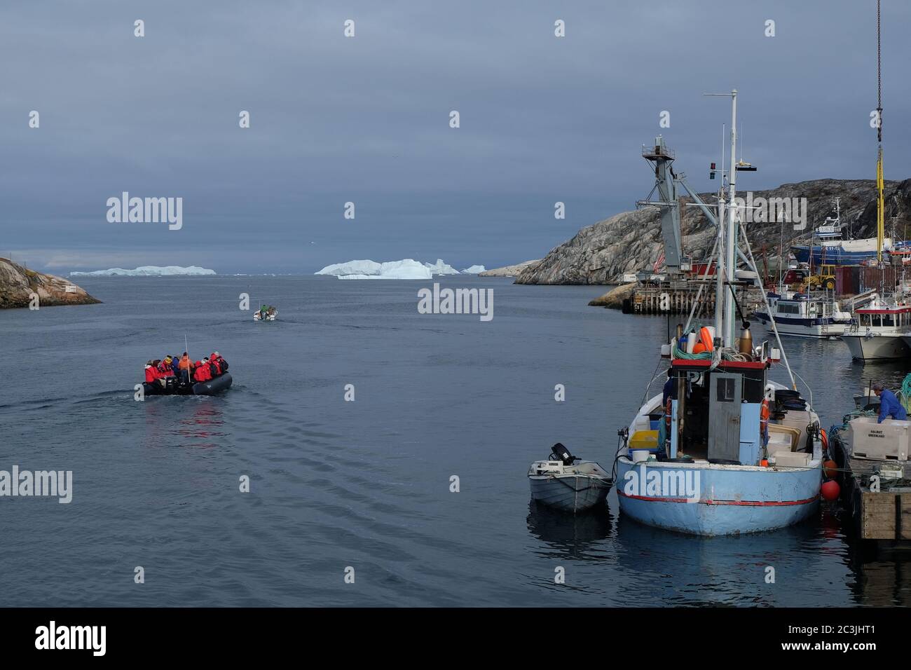 Un petit bateau de pêche Greenlandic décharge sa prise tandis qu'un groupe de touristes vêtus de rouge se dirige vers Disco Bay pour observer les baleines et naviguer autour d'énormes icebergs. Banque D'Images