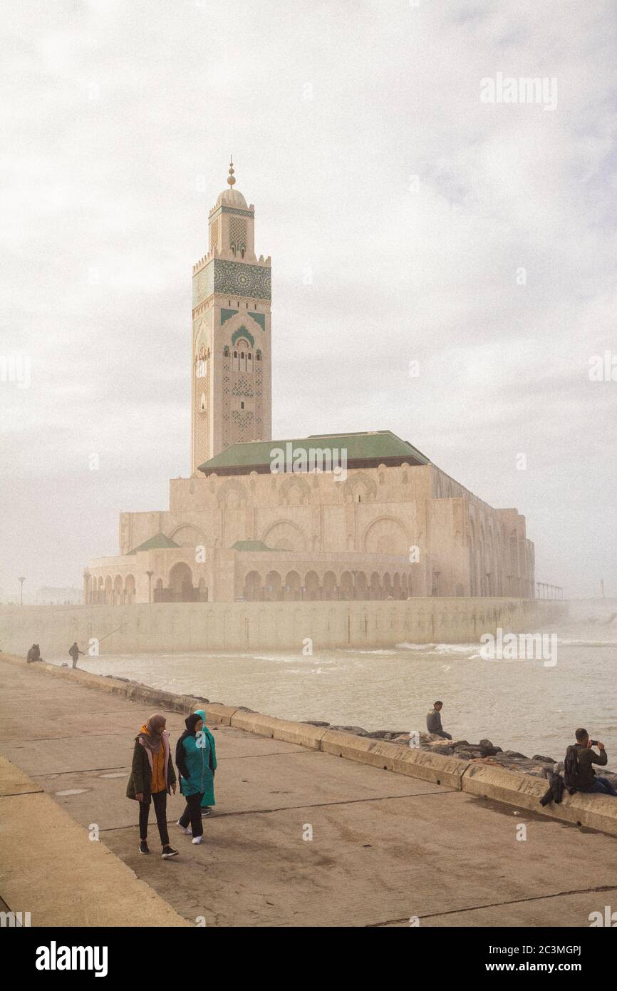 Un groupe de personnes marchant et assis autour de la mosquée Hassan II à Casablanca, au Maroc, pendant un après-midi brumeux, profitant de la vue sur l'océan Banque D'Images