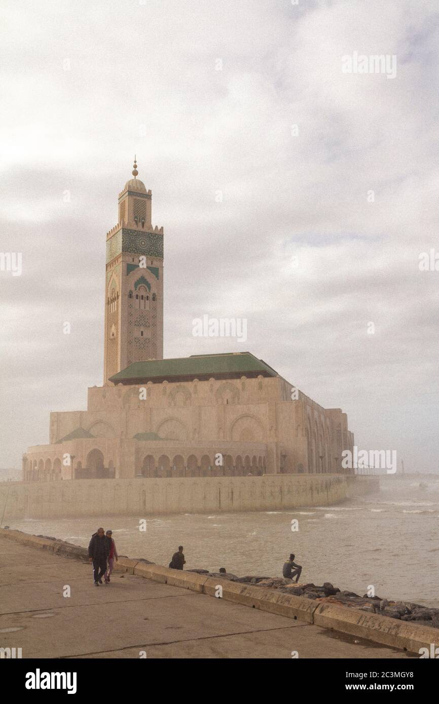 Un groupe de personnes marchant et assis autour de la mosquée Hassan II à Casablanca, au Maroc, pendant un après-midi brumeux, profitant de la vue sur l'océan Banque D'Images