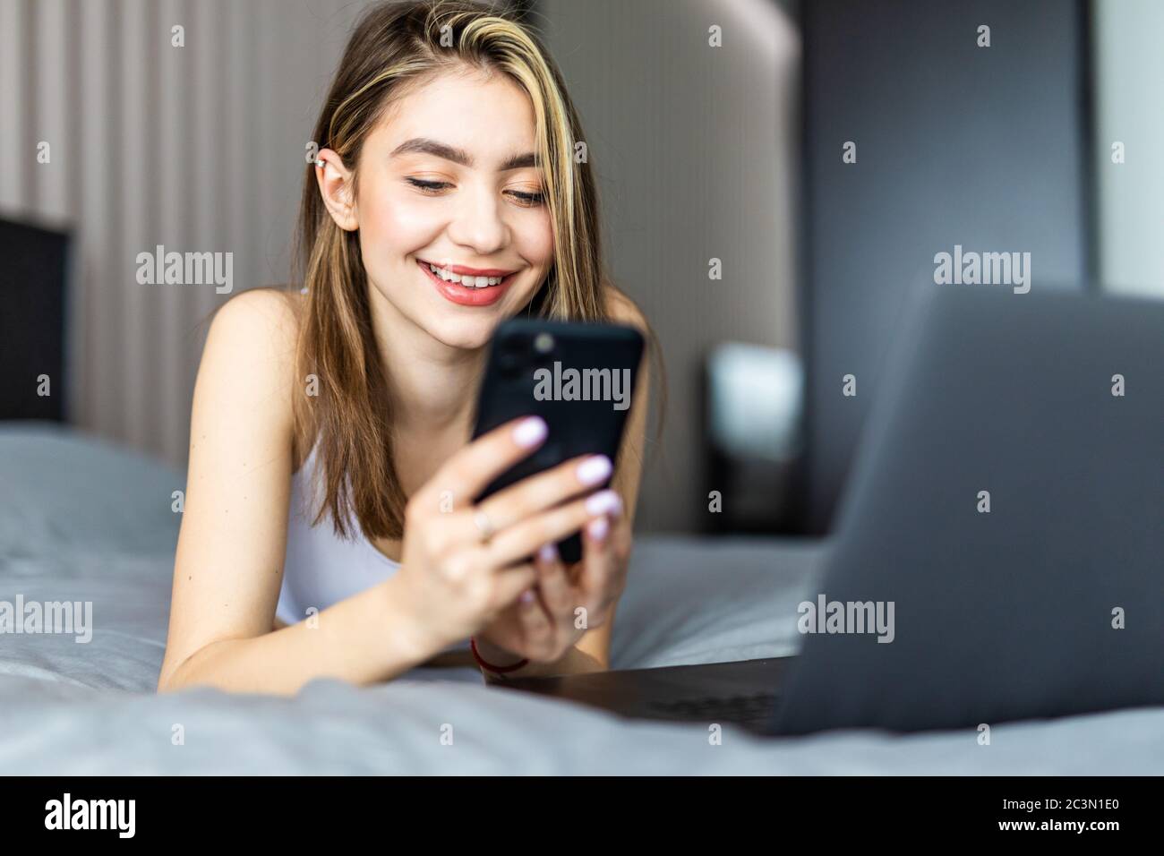 Une belle femme souriante, couchée sur le lit près de l'ordinateur portable et tapant un message sur son téléphone. Banque D'Images