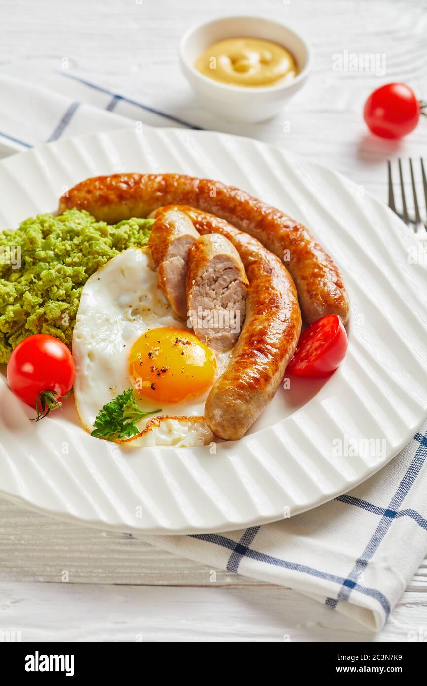 Assiette blanche avec de longues saucisses de porc, œufs frits côté ensoleillé, tomates cerises, purée de pois verts sur un fond en bois clair avec moutarde et couverts Banque D'Images