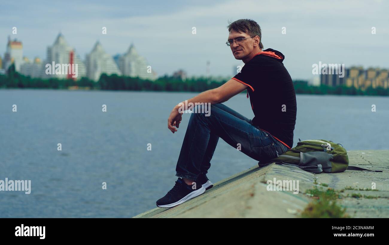 Un homme avec des lunettes près de la rivière. Portrait d'un homme en lunettes et vêtements décontractés, assis près de la rivière par une journée nuageux Banque D'Images