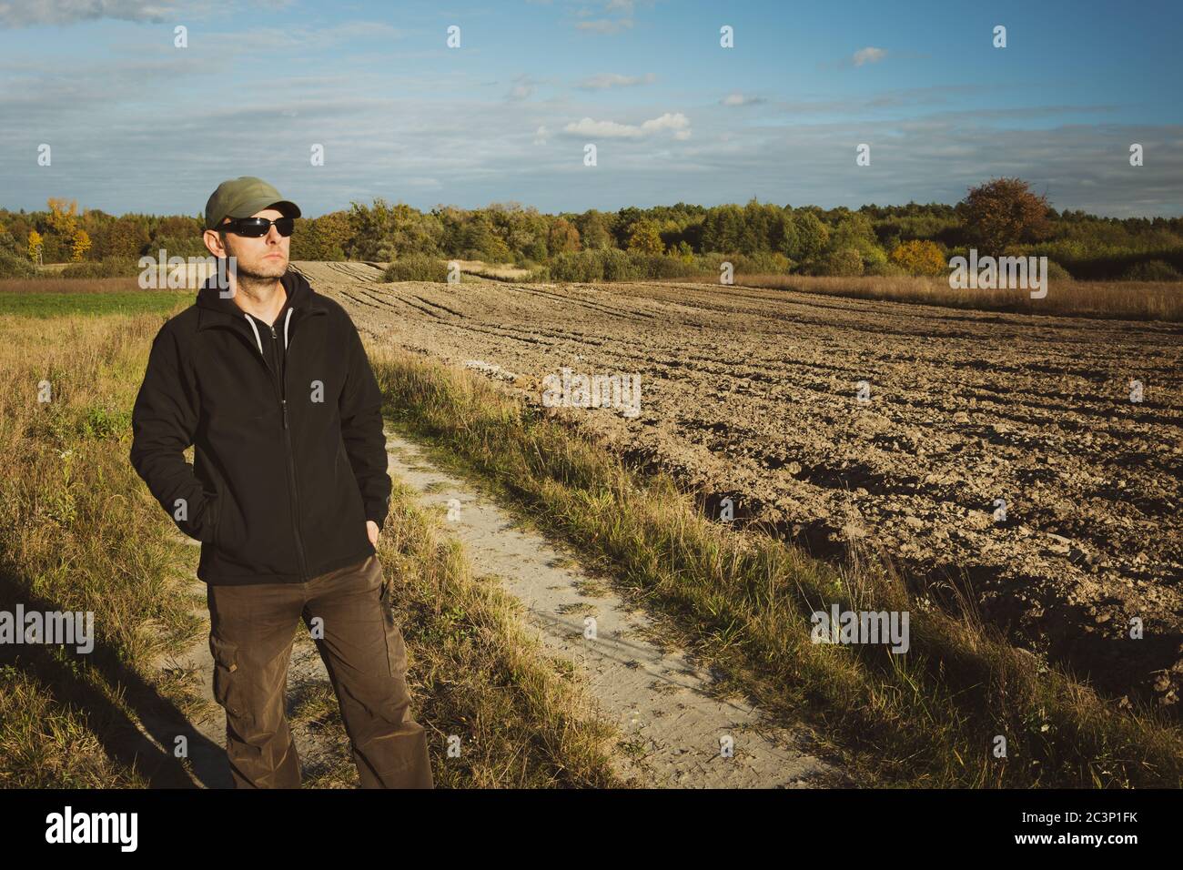 Un homme en lunettes de soleil debout près d'un champ labouré, vue rurale ensoleillée Banque D'Images