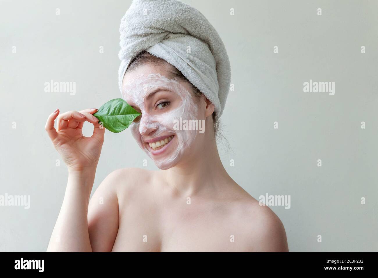 Portrait de beauté minime fille en serviette sur la tête application du masque nourrissant blanc ou crème sur le visage, feuille verte dans le fond blanc isolé main. Soin de la peau nettoyage éco-biologique cosmétique spa concept Banque D'Images