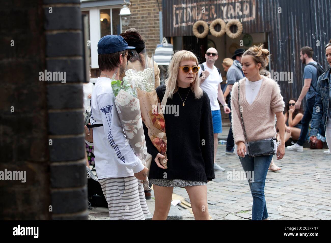 London, UK - 17 juillet 2019, Columbia Road Flower Market, une fille blonde sérieuse en lunettes de soleil, chandail noir, jupe courte en Jean avec un bouquet de Banque D'Images