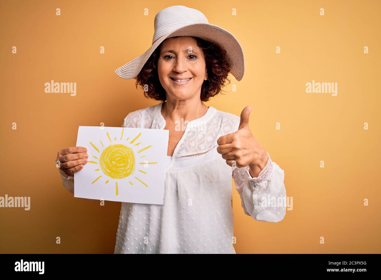 Femme maurique d'âge moyen en vacances tenant bunner avec image de soleil sur fond jaune heureux avec grand sourire faisant signe ok, pouce vers le haut avec les doigts, sauf Banque D'Images