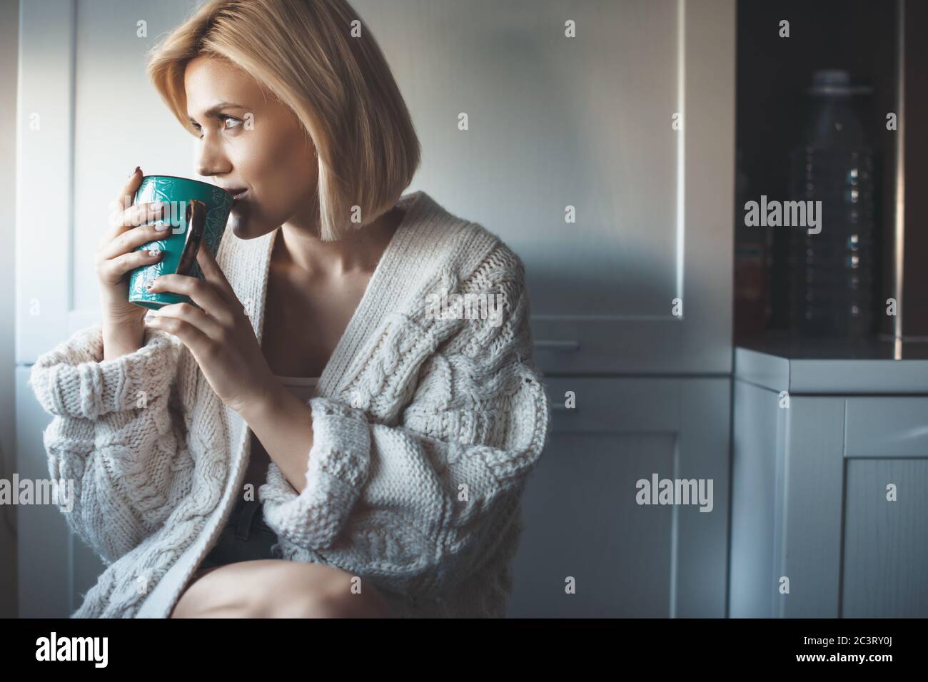 Belle femme blonde avec pull en tricot boire un thé près de la fenêtre et sourire regardant quelque part Banque D'Images