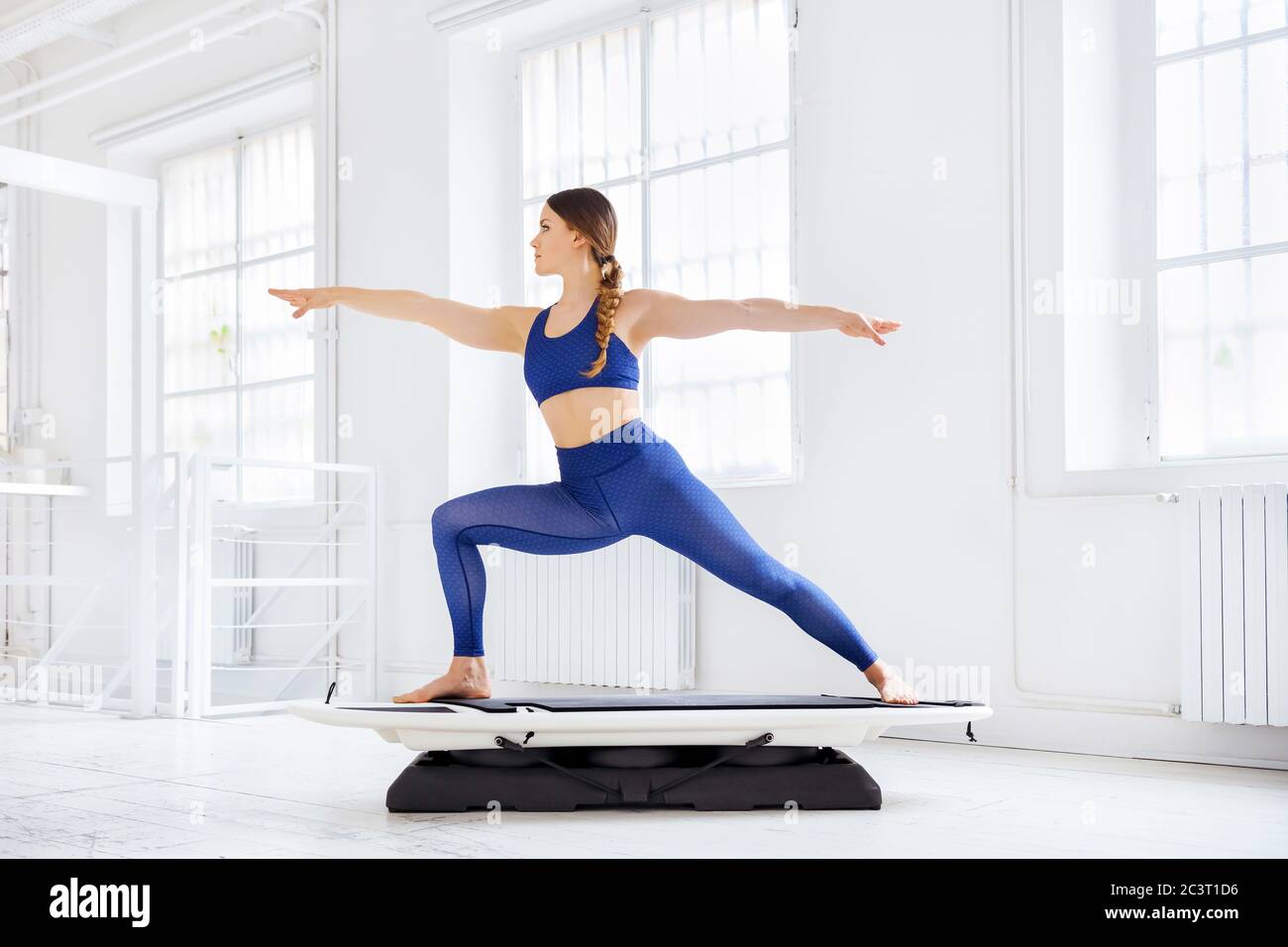 Femme effectuant une posture de yoga Warrior 2 ou s'étire sur un surf de forme physique ou de planche dans une vue latérale dans une salle de gym de haut-clé dans un concept de santé et de forme physique Banque D'Images