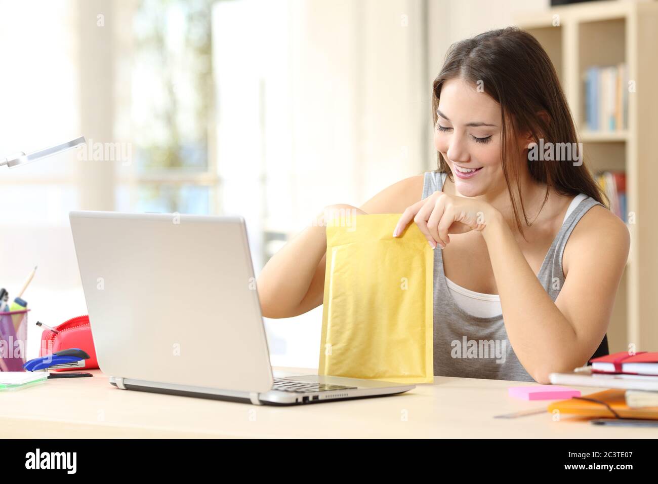 Bonne étudiante femme avec ordinateur portable ouvrant enveloppe rembourrée assise sur un bureau à la maison Banque D'Images