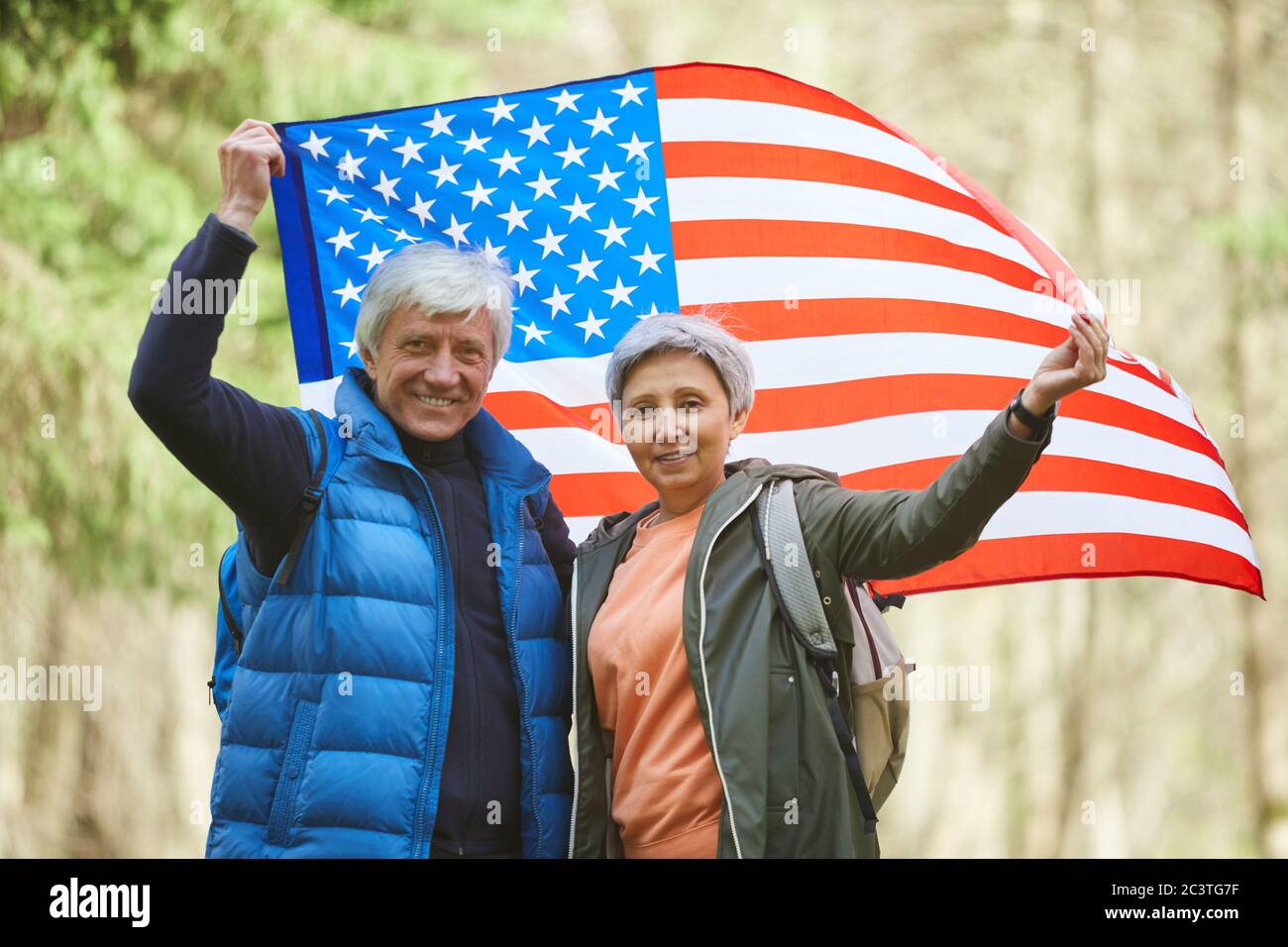 Portrait de couple senior actif tenant le drapeau américain et souriant à l'appareil photo tout en appréciant la randonnée dans la forêt, espace de copie Banque D'Images