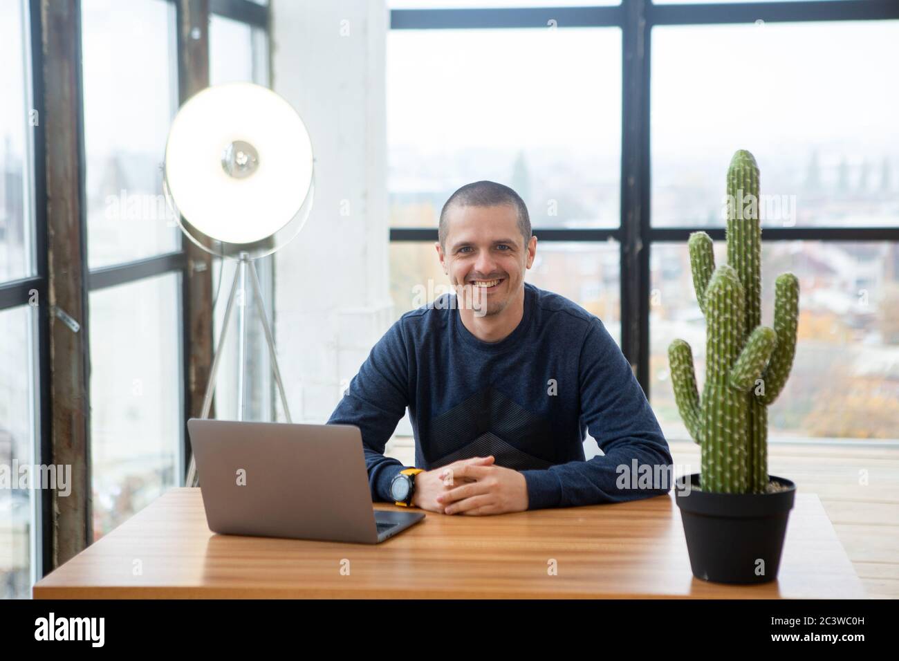 Homme adulte souriant regardant dans l'appareil photo tout en étant assis au bureau avec ordinateur portable. Banque D'Images