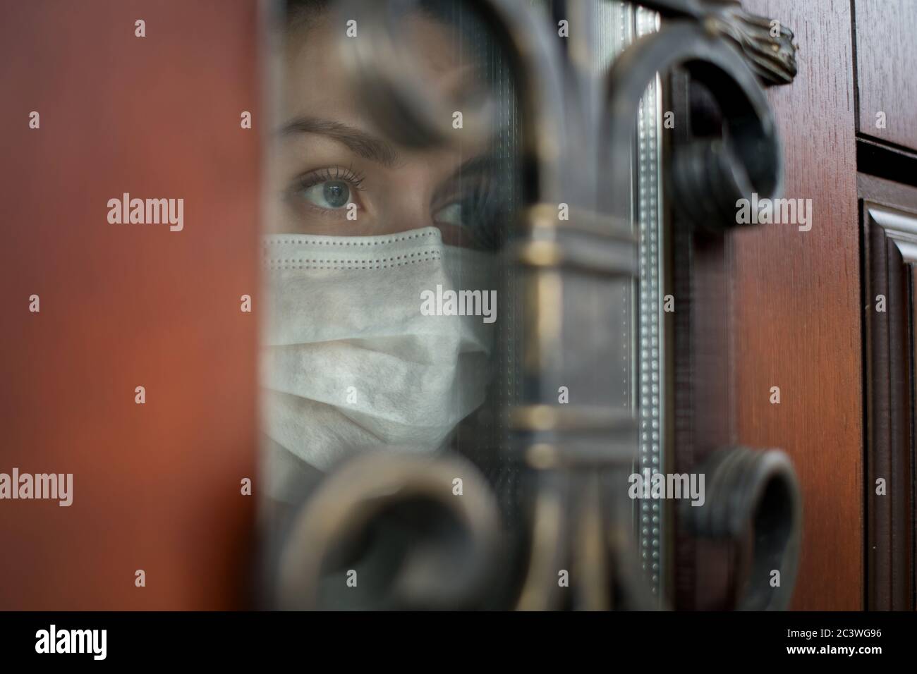 Une jeune femme dans un masque médical regarde à travers le porte d'entrée de la rue Banque D'Images