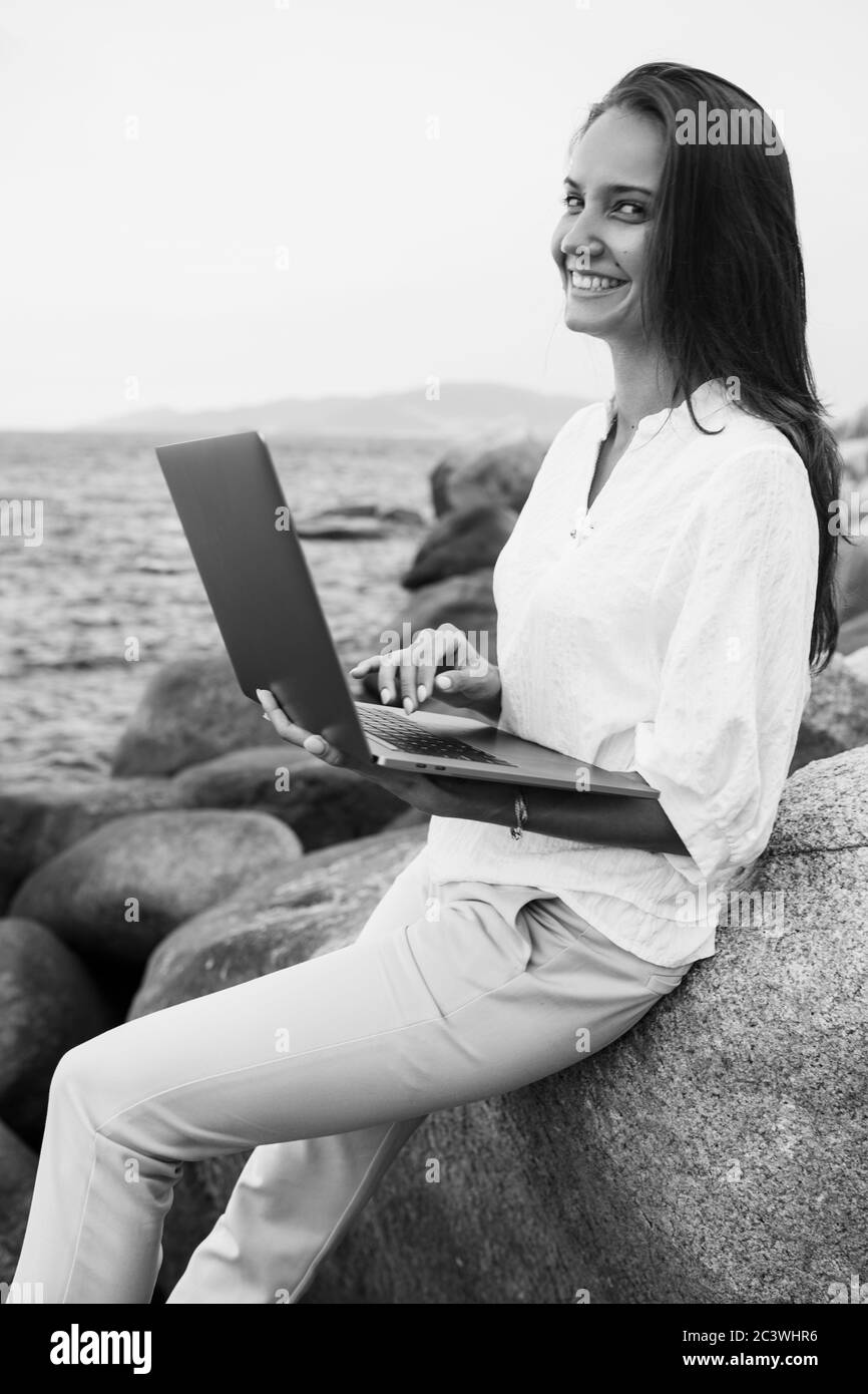 Photo noir et blanc Jeune fille souriante et réussie travaillant avec un ordinateur portable, assise sur un rivage rocheux. Femme d'affaires travaillant à distance pendant Banque D'Images