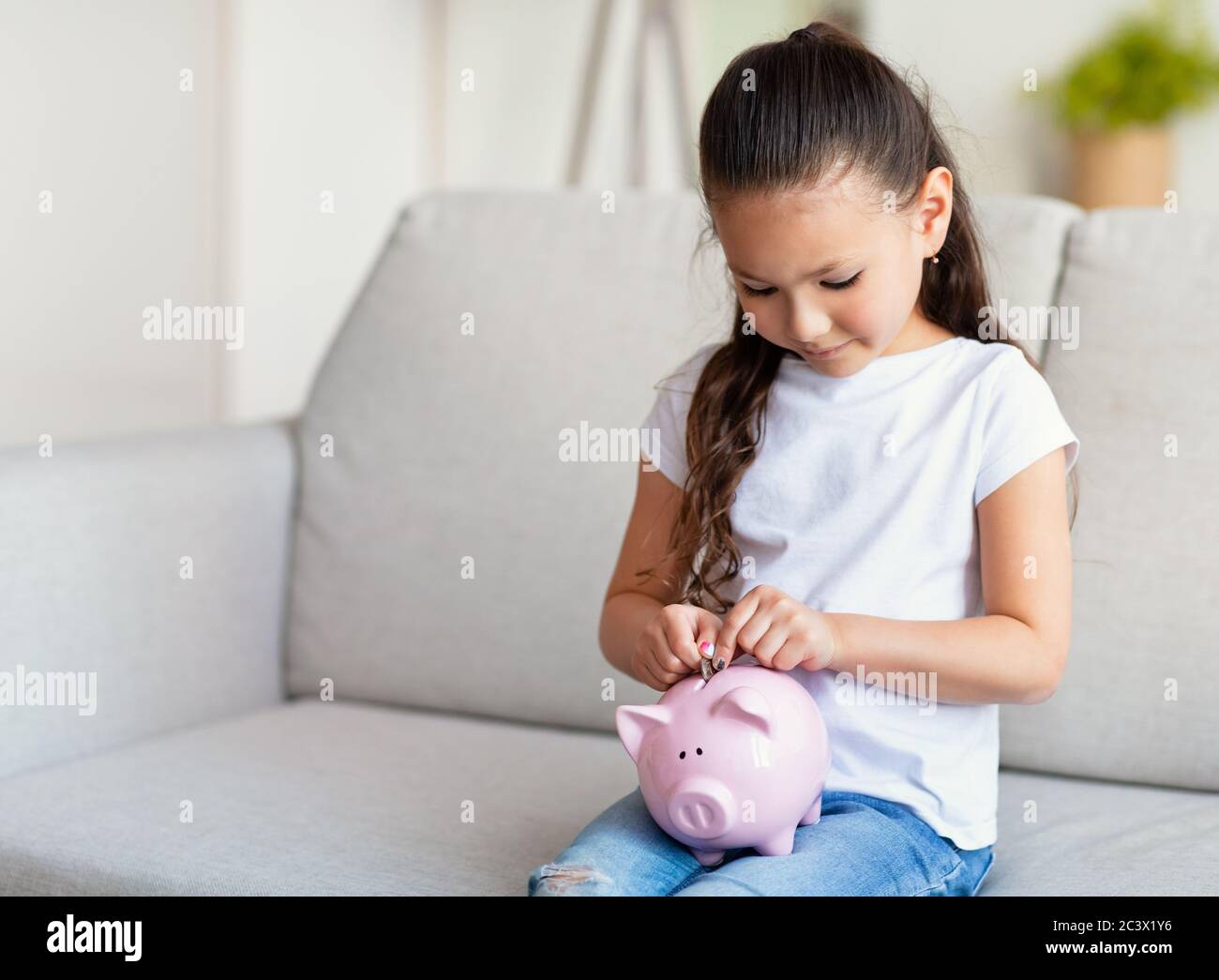 Fille mettant pièce dans Piggybank assis sur canapé à la maison Banque D'Images