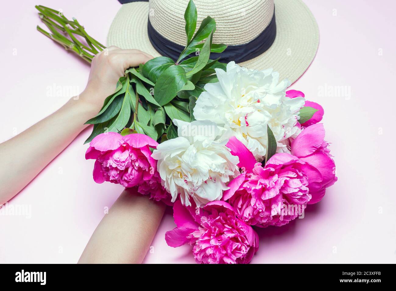 Mélanger le bouquet de pivoines et le chapeau de paille avec un ruban noir dans les mains femelles isolées sur fond rose. Composition de la pose à plat, gros plan, vue de dessus Banque D'Images
