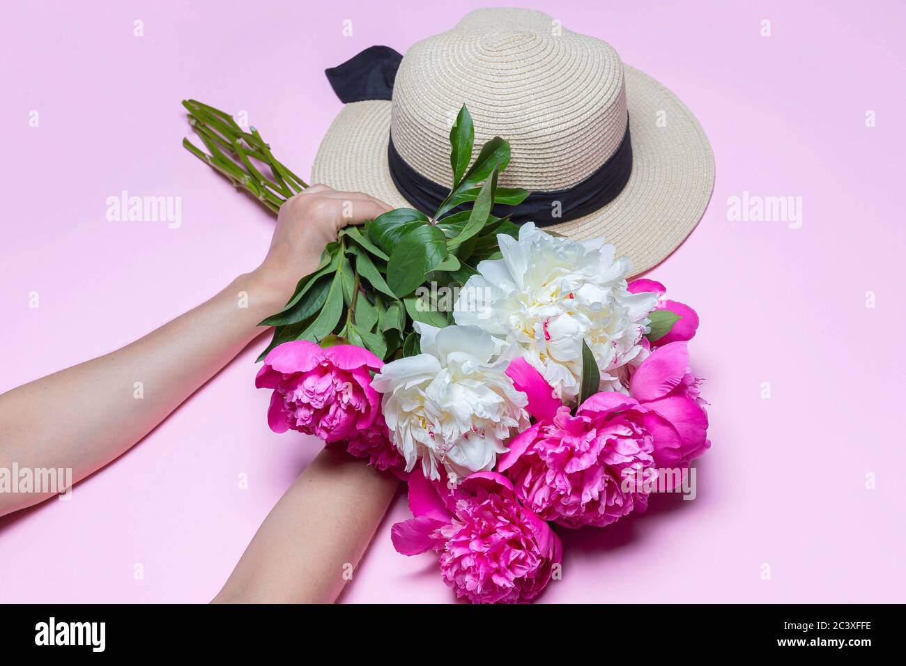 Chapeau de paille avec ruban noir et bouquet de pivoines dans les mains de femmes isolées sur fond rose. Composition de la pose à plat, vue de dessus, espace de copie Banque D'Images