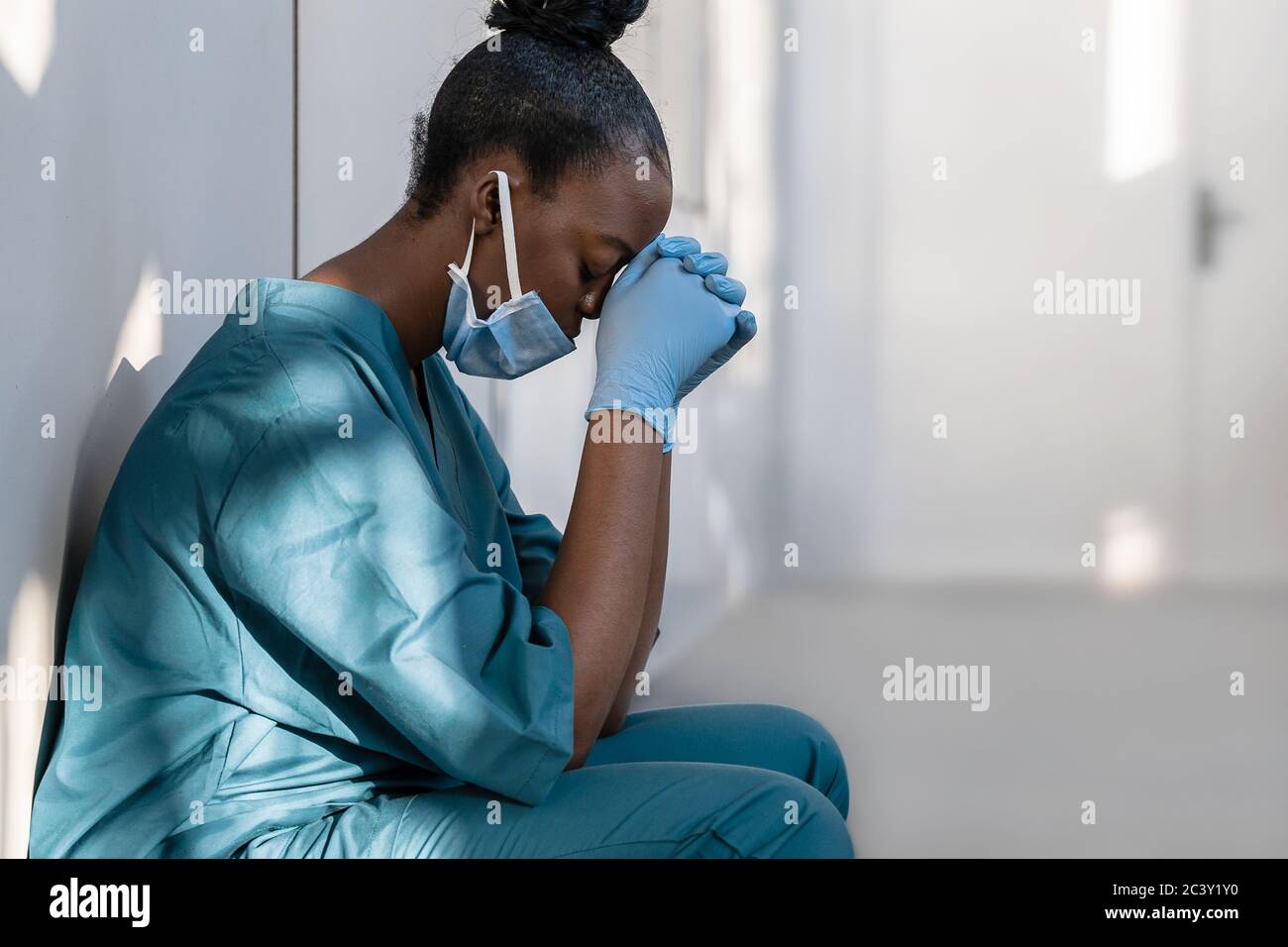 Une infirmière africaine fatiguée et dépressive portant un masque facial est assise sur le sol de l'hôpital. Banque D'Images