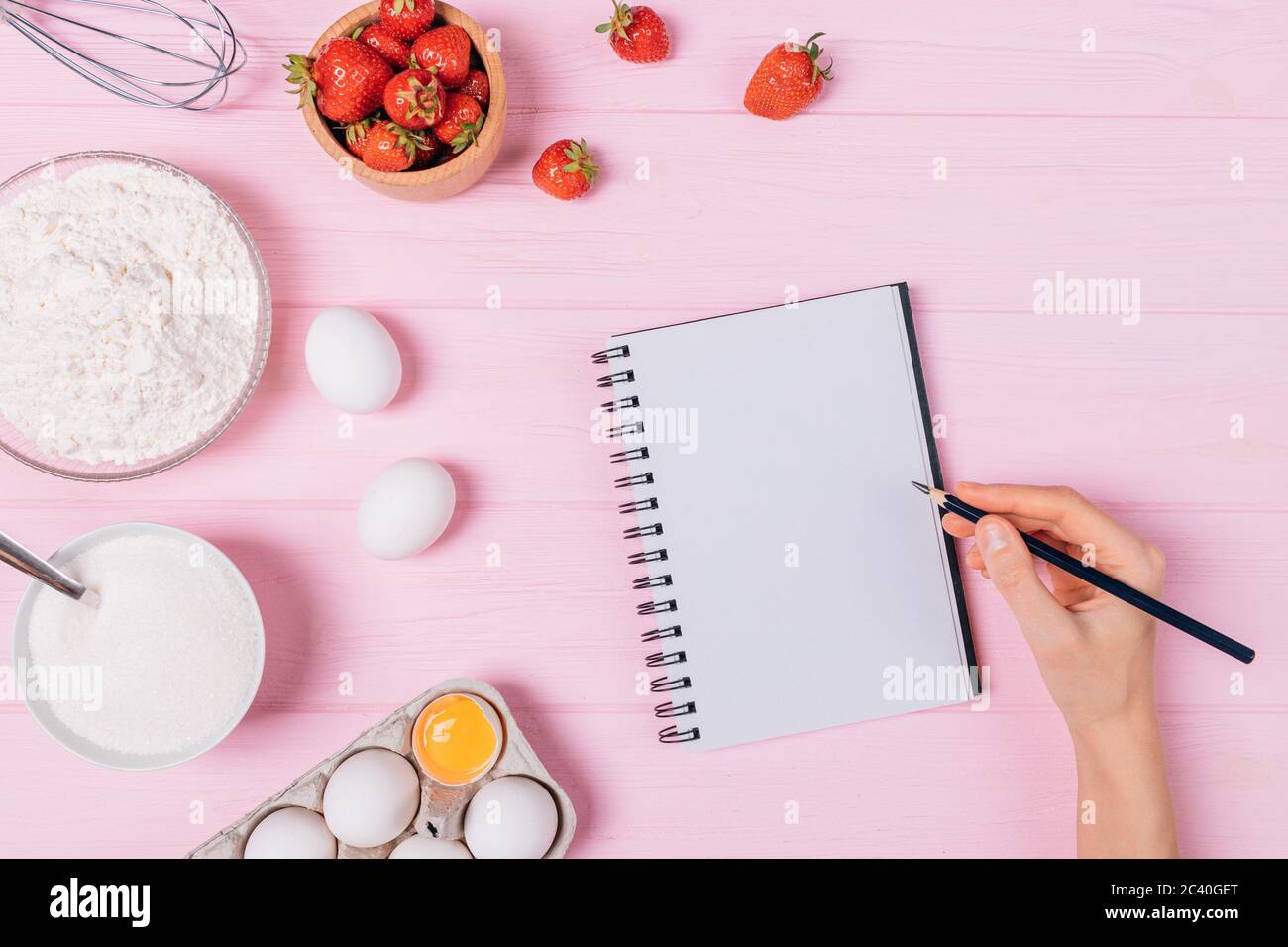 Recette de tarte aux fraises maison, écriture manuelle de femme dans un carnet vide à côté des ingrédients fruits frais, oeufs, farine et sucre sur table rose, Fla Banque D'Images